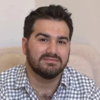 A man with dark hair and a beard, wearing a checkered shirt, sitting indoors against a light background.