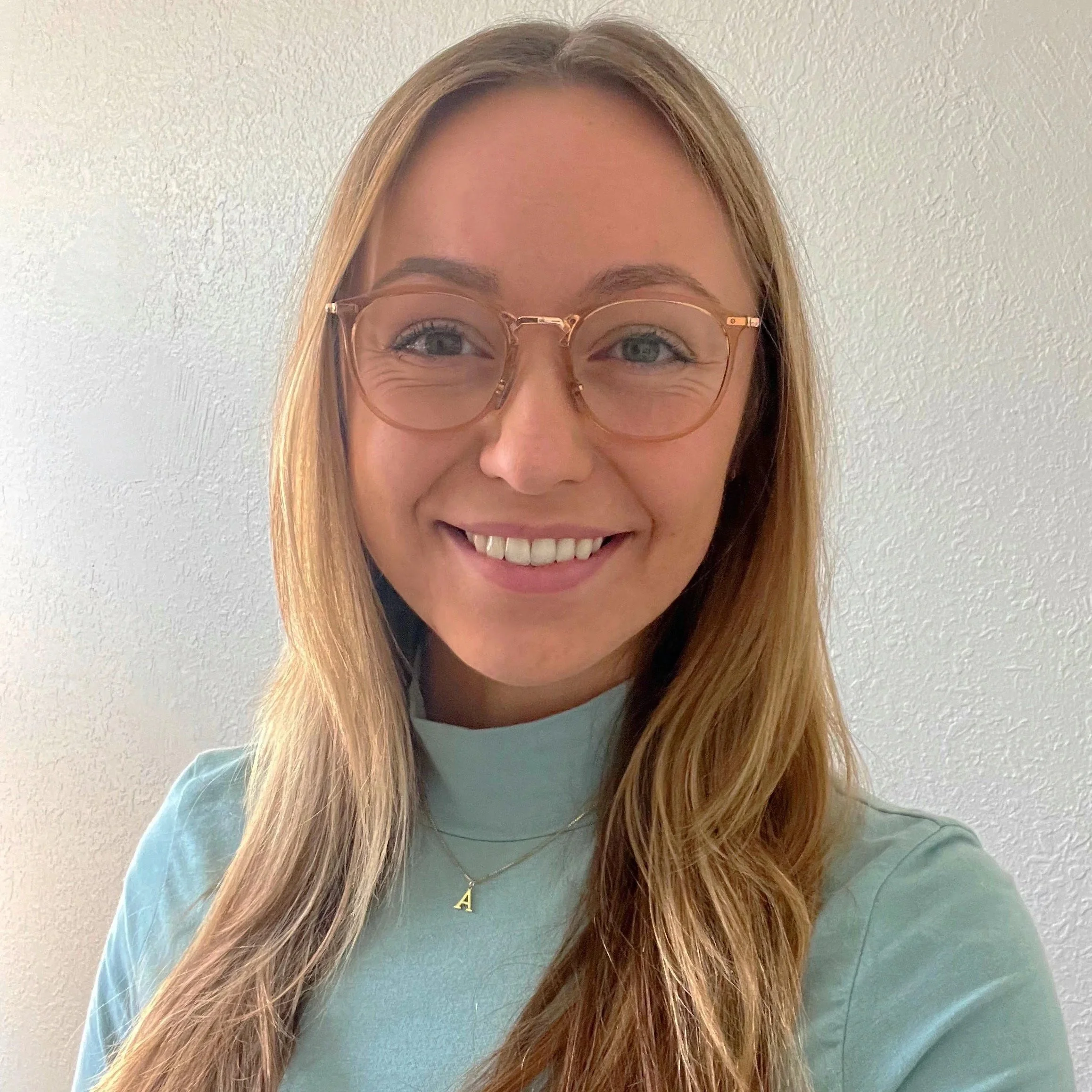 A young woman with long, light brown hair and glasses smiling at the camera, wearing a light blue top and a gold necklace with the letter 'A' against a white textured wall.