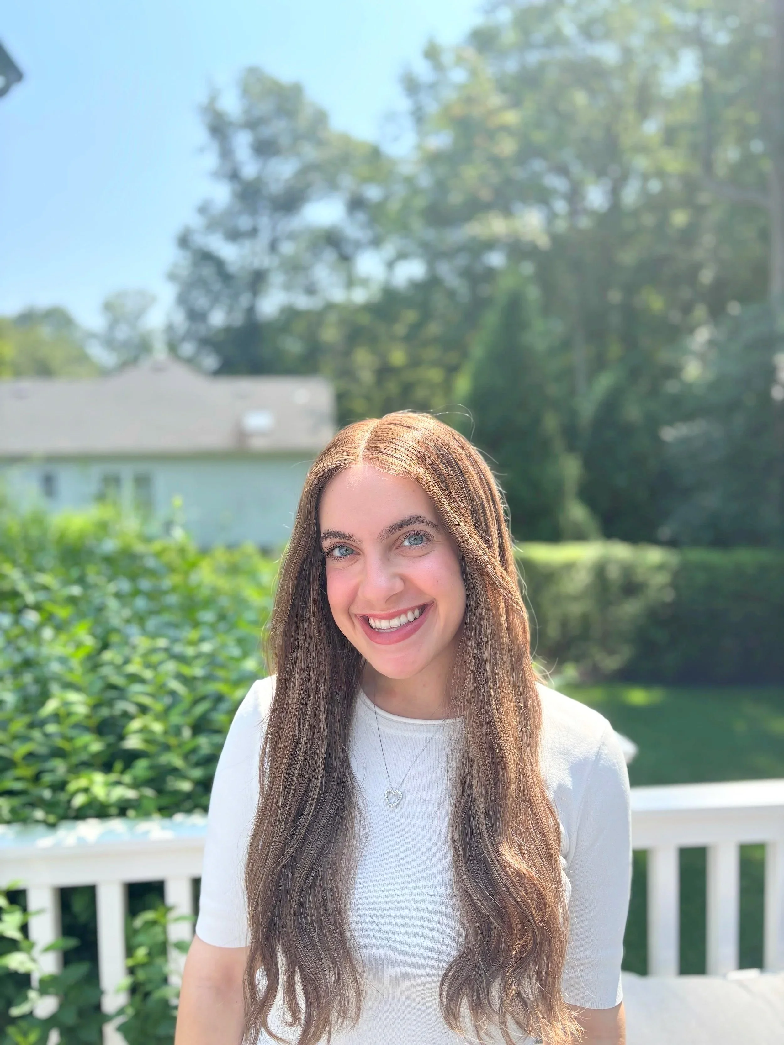 A young woman with long, wavy brown hair and blue eyes smiling outdoors on a sunny day, wearing a white shirt and a necklace with a heart pendant.