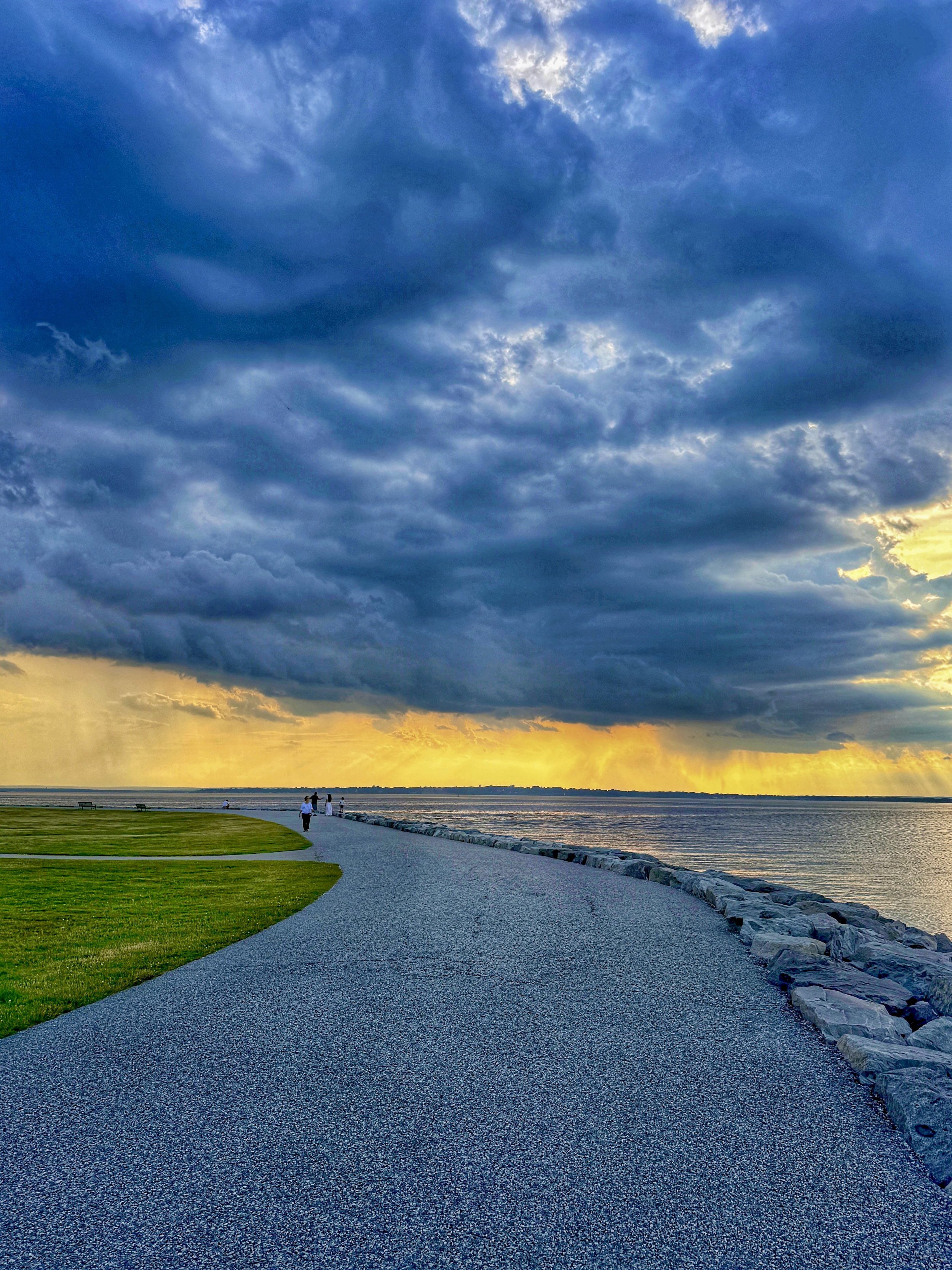Gold and orange golden hour sunset rays and moody gray clouds over Narragansett Bay as you walk  along the shoreline at Colt State Park in Bristol, Rhode Island.