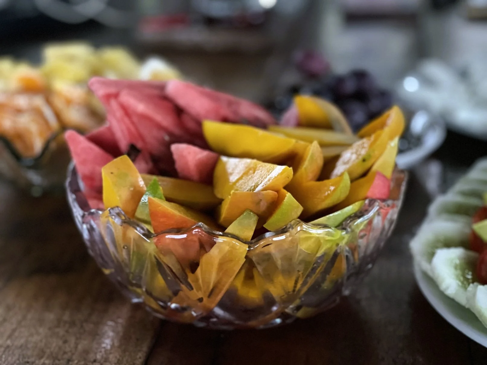 A glass bowl of mixed fruit including watermelon, mango, and melon on a wooden table with more dishes in the background, served at Casa del Maestro ayahuasca center in Peru.