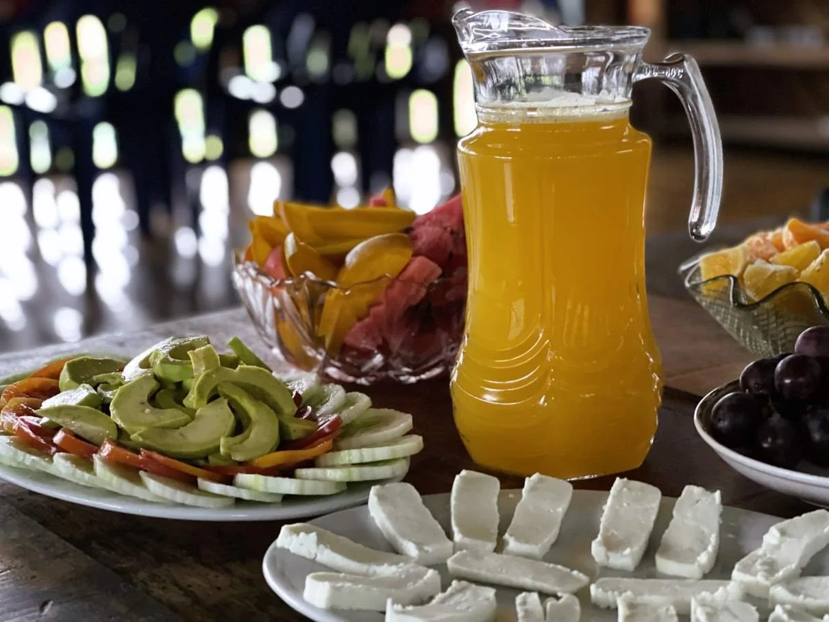 A pitcher of maracuya juice, a plate of sliced avocado and vegetables, a bowl of fruit salad, and sliced white cheese on a wooden table at an ayahuasca retreat in Peru.
