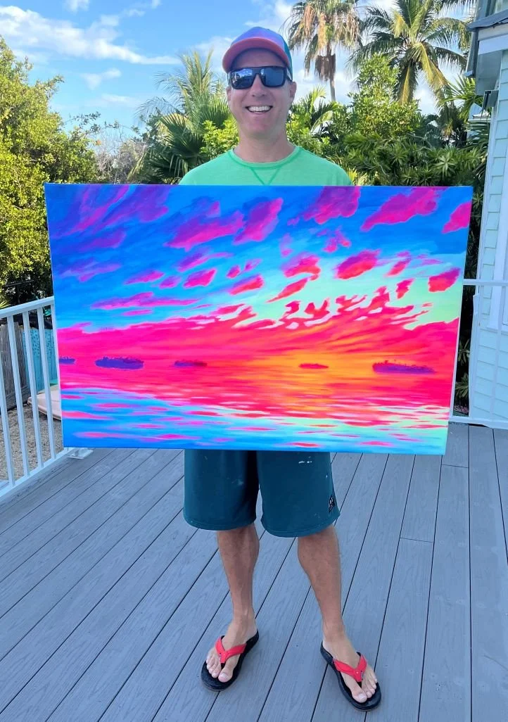 A man wearing sunglasses, a colorful cap, a green shirt, and flip-flops is holding a painting of a vibrant sunset over the water with pink, purple, orange, and blue clouds and reflections.