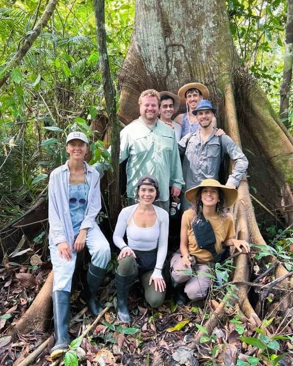 Group of ayahuasca retreat guests posing together in a jungle in Peru, surrounded by dense greenery and a large lupuna tree.