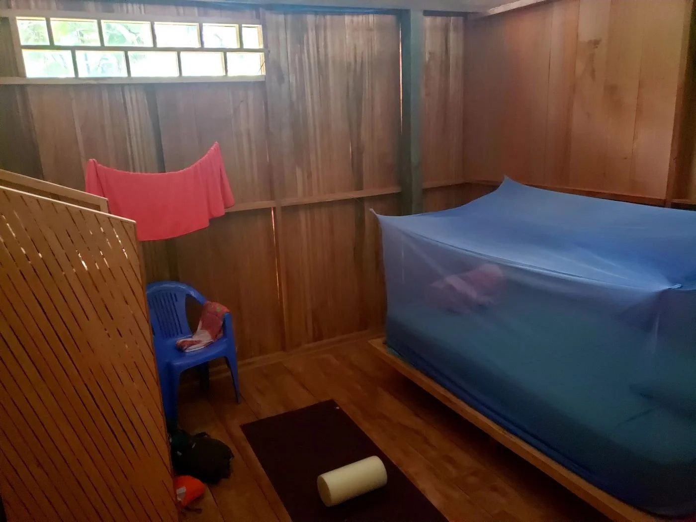 A guest bedroom with wooden walls and floors, a bed covered with a blue mosquito net, a blue plastic chair, a black mat with a white rolled towel, a small window with glass blocks at Casa del Maestro ayahuasca retreat center.