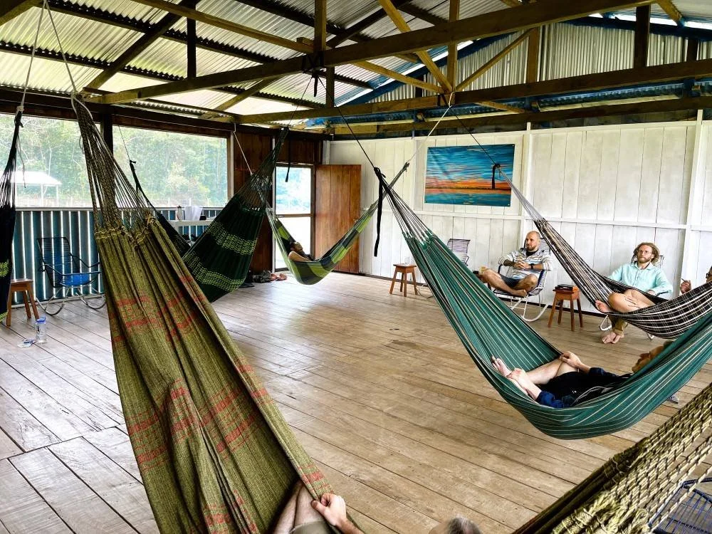 Retreat guests relaxing in hammocks inside a rustic wooden shelter with a metal roof and wooden floor, at Casa del Maestro ayahuasca center.
