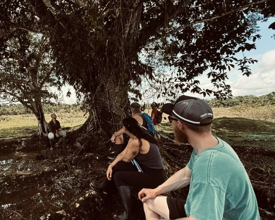 A group of people sit near a tree in an open nature setting at an ayahuasca retreat center in Peru.