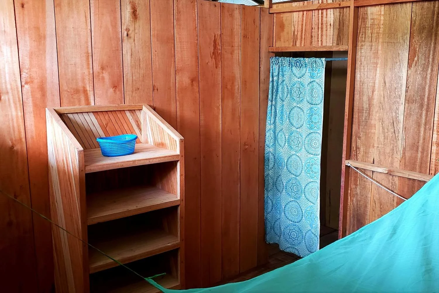 Wood-paneled room with a small wooden bookshelf, a blue bucket on top, and a doorway with a blue patterned curtain at Casa del Maestro ayahuasca retreat center in Peru.