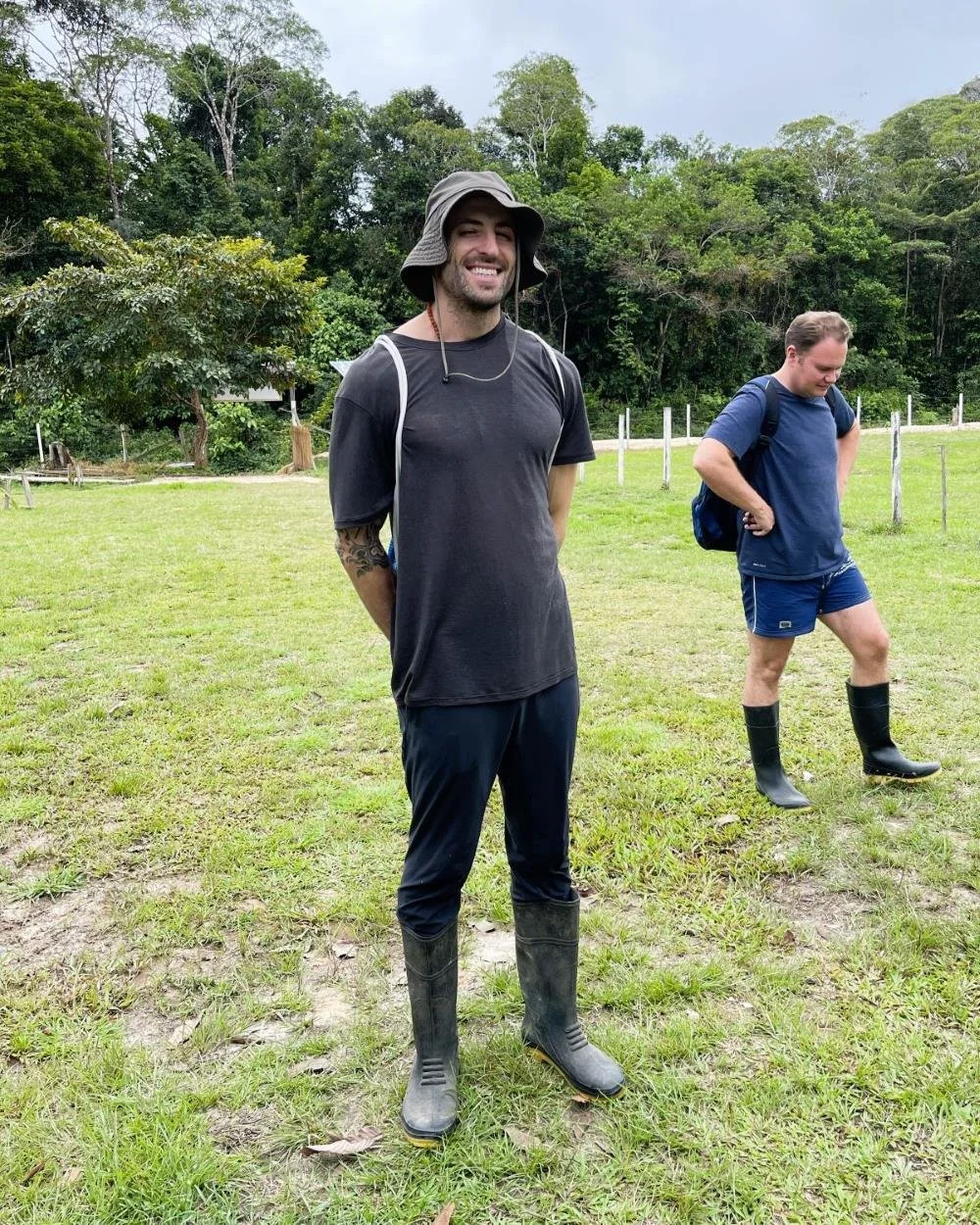 Two men standing in grassy field with trees in background. The man in the foreground is smiling, wearing a wide-brimmed hat, black t-shirt, rubber boots, and has a tattoo on his right arm. The man in the background has short hair, is looking down, and is dressed in a blue t-shirt, shorts, rubber boots, and is carrying a backpack.