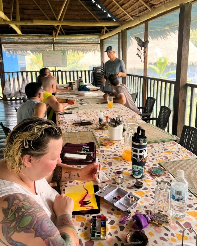 A group of ayahuasca and art retreat participants seated around a large dining table in a tropical setting, coloring and drawing in notebooks.