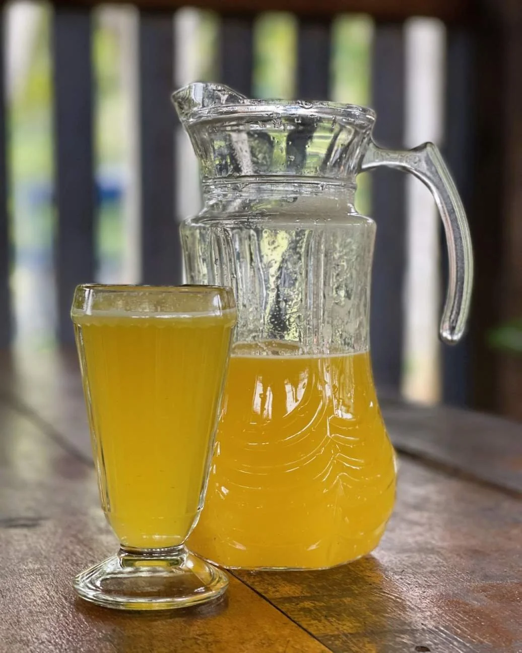 A glass pitcher and a tall glass filled with maracuya juice placed on a wooden table, with a blurred outdoor background of a jungle retreat center.