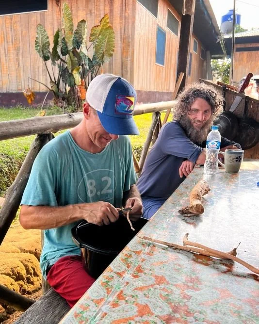 Two men sitting at an outdoor table in a rustic setting, scraping uchu sanango roots, with a wooden building and tropical plants in the background.