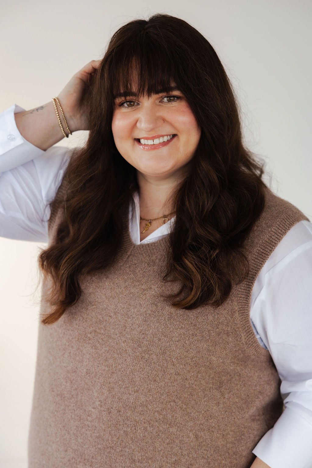 A woman with long, dark, wavy hair and bangs smiling, wearing a white shirt under a brown vest, with jewelry, against a plain light background.