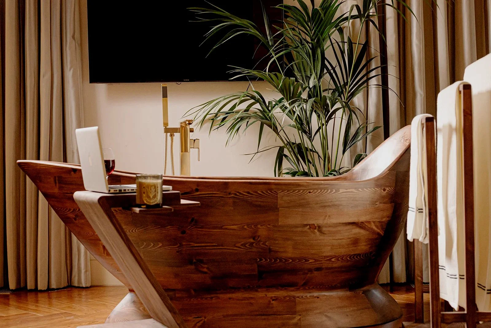 Elegant bathroom with a wooden bathtub, large green plant, television, and towels hanging on a wooden rack.