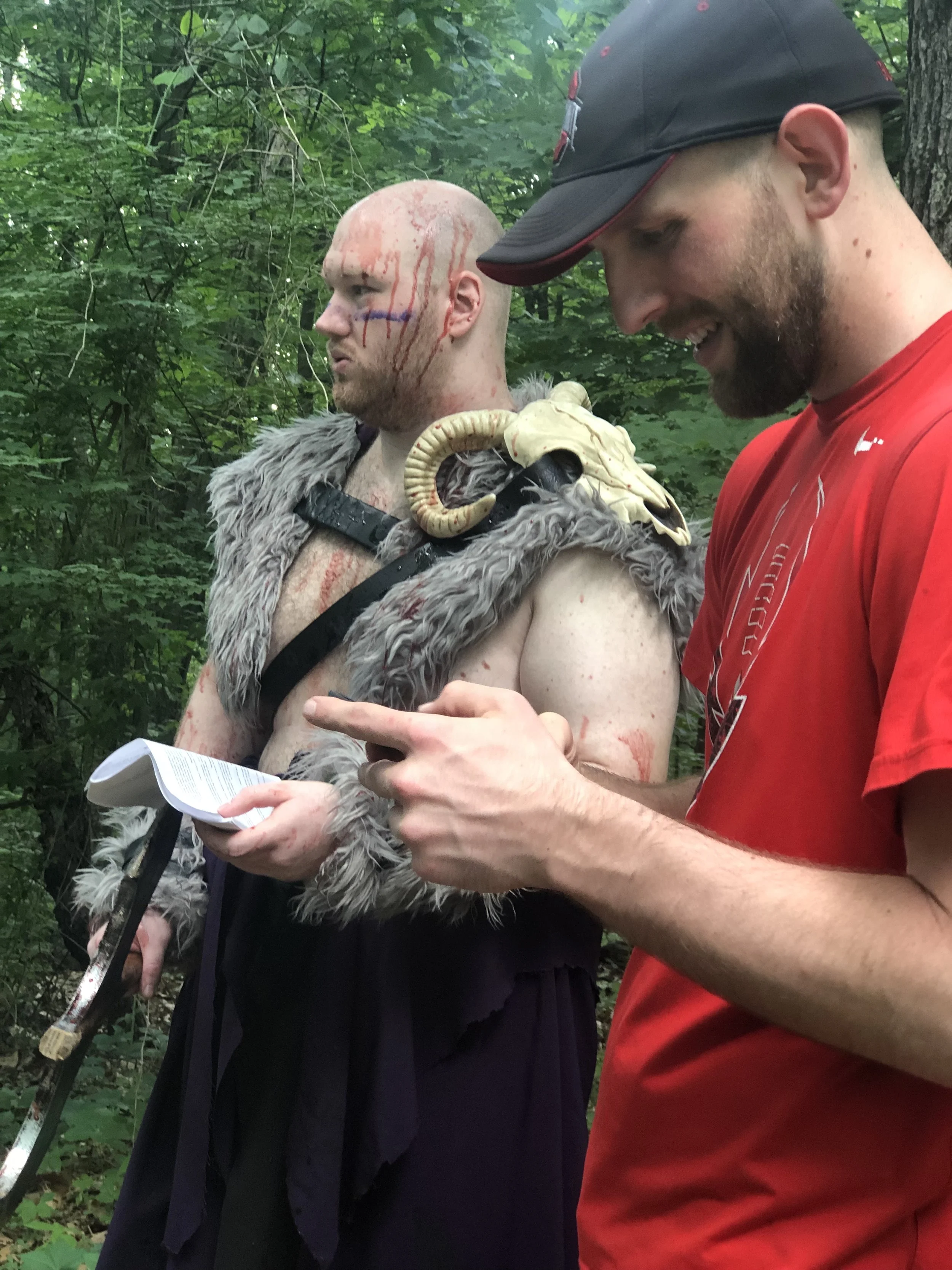 Two men in a forested area, one of whom is dressed in costume with a fur cloak, a ram skull shoulder piece, and face paint, while the other wears a red shirt and a black cap, and they are looking at a piece of paper.