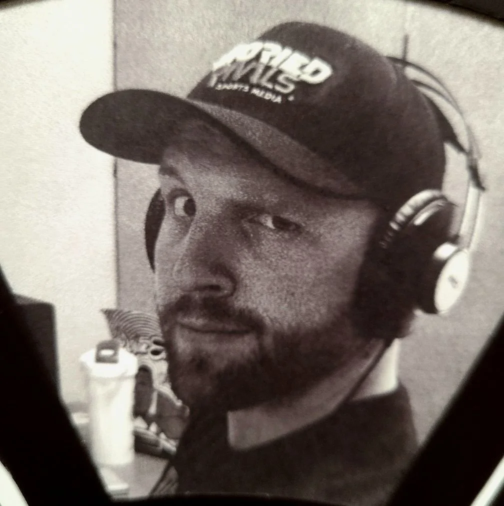 A black-and-white photo of a man wearing a baseball cap and headphones, looking at the camera.