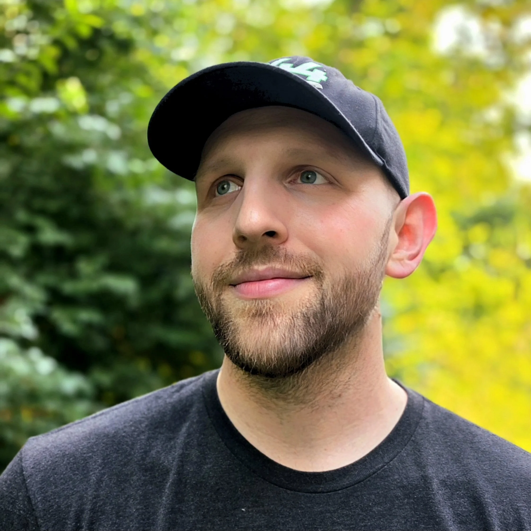 Close-up of a young man's face with blue eyes, wearing a dark cap and gray T-shirt, looking slightly to the side, outdoors with green foliage in the background.