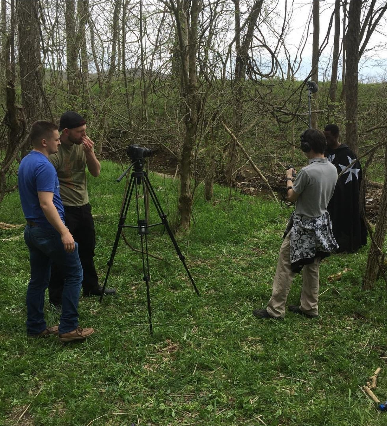 A group of five people in a wooded area, with some operating filming equipment and others observing. One person is looking through binoculars, and there is a camera on a tripod pointed into the trees.
