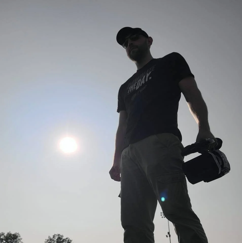 Silhouette of a man wearing a cap and sunglasses, holding a camera, standing outdoors against a bright sun in the sky.