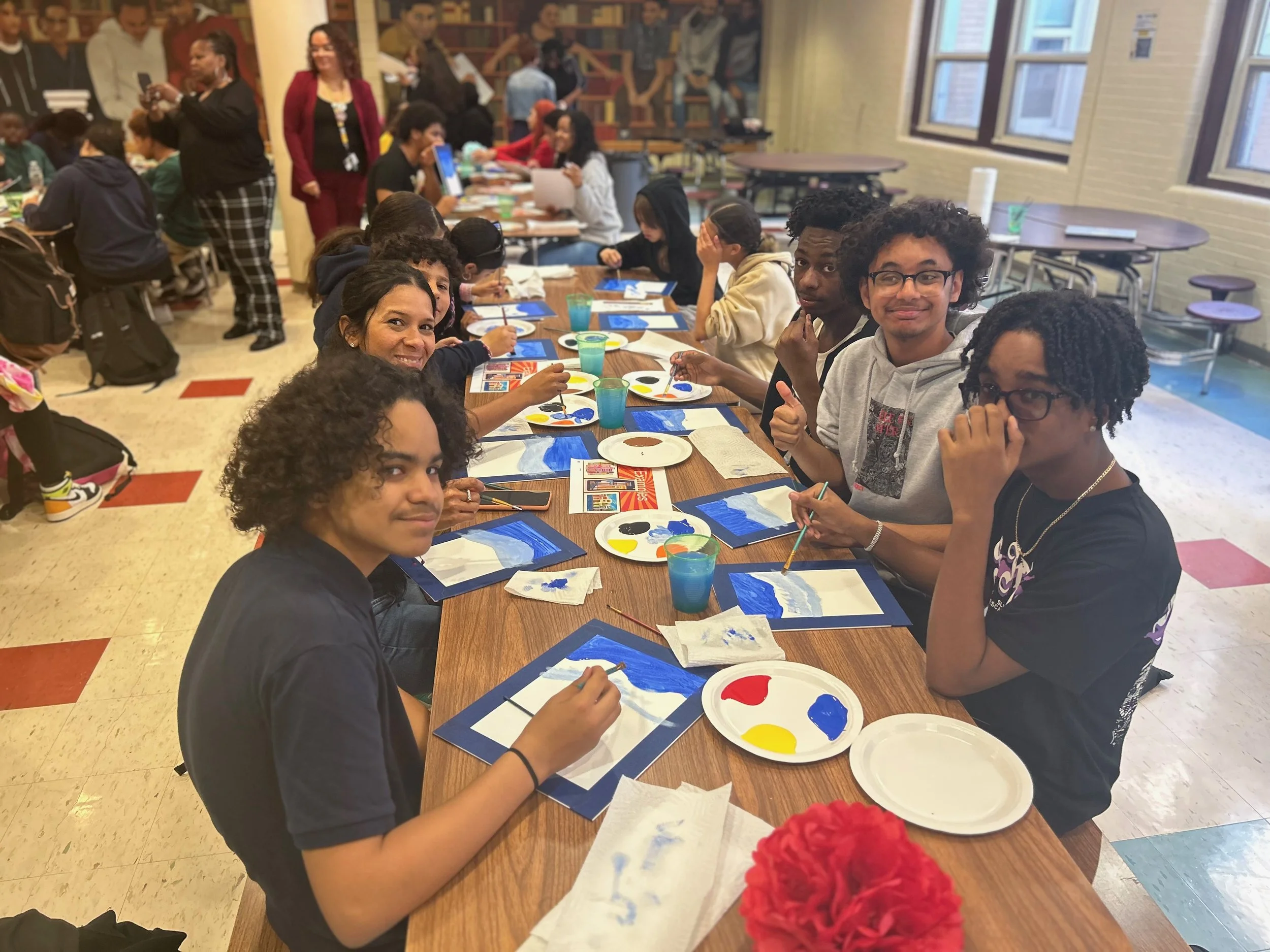 Group of young students and teachers at a classroom table painting with blue, yellow, black, and white paints for an art project.