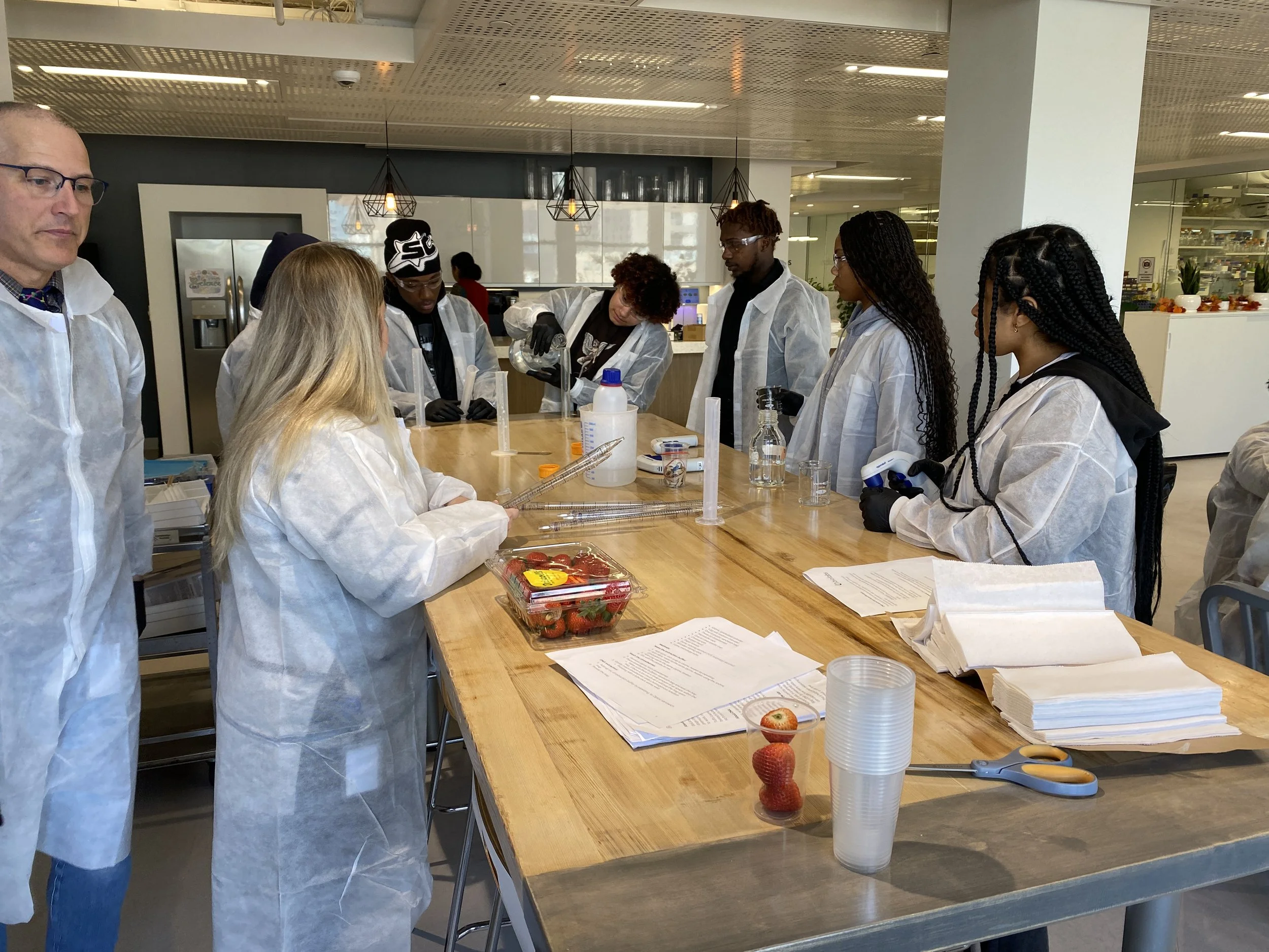 People in lab coats conducting a scientific experiment or demonstration in a laboratory setting with equipment, papers, strawberries, and a plastic container on the table.