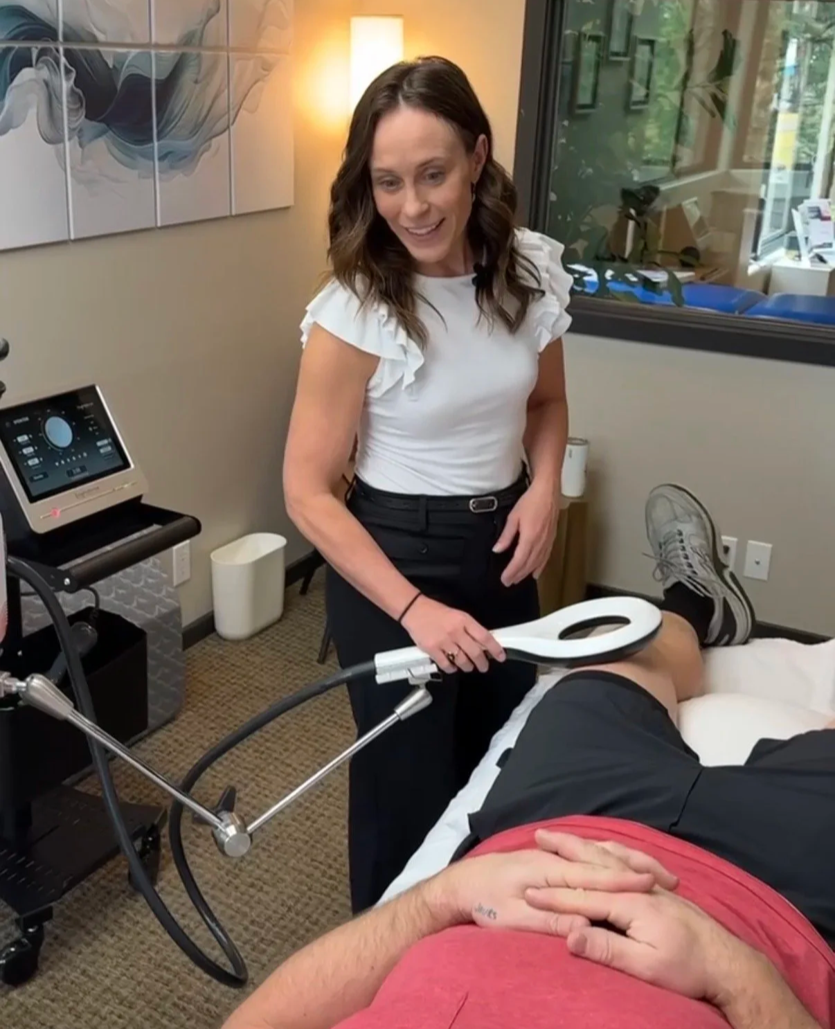 Maria Delliveneri of Remedy Sports Medicine utilizing EMTT on a patient's arthritic knee. They are in a cozy treatment room, the patient is laying on their back on a treatment table in their gym clothes.