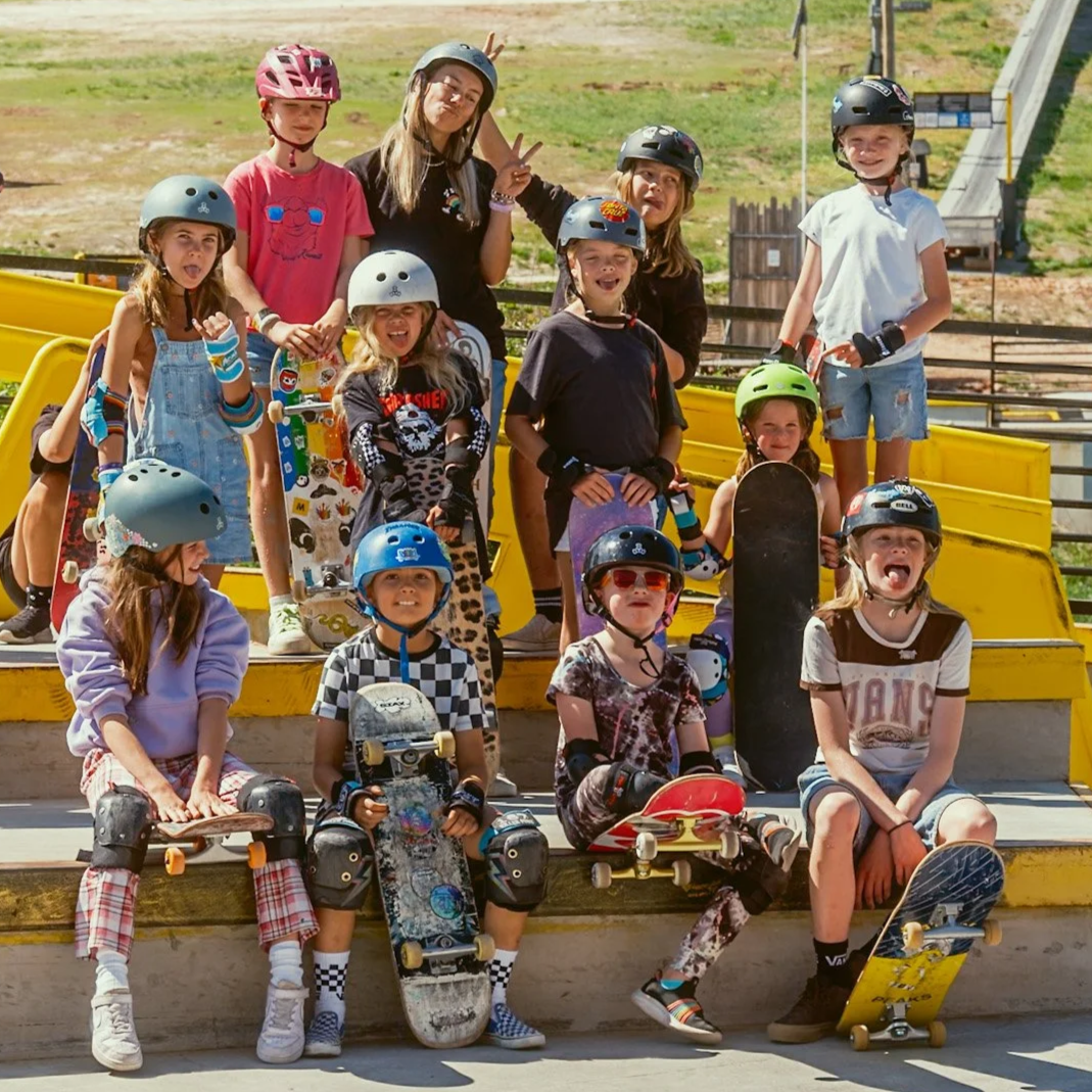 Group of children and a teenage girl at skate park, all wearing helmets, some with skateboards, posing for photo on yellow stairs.