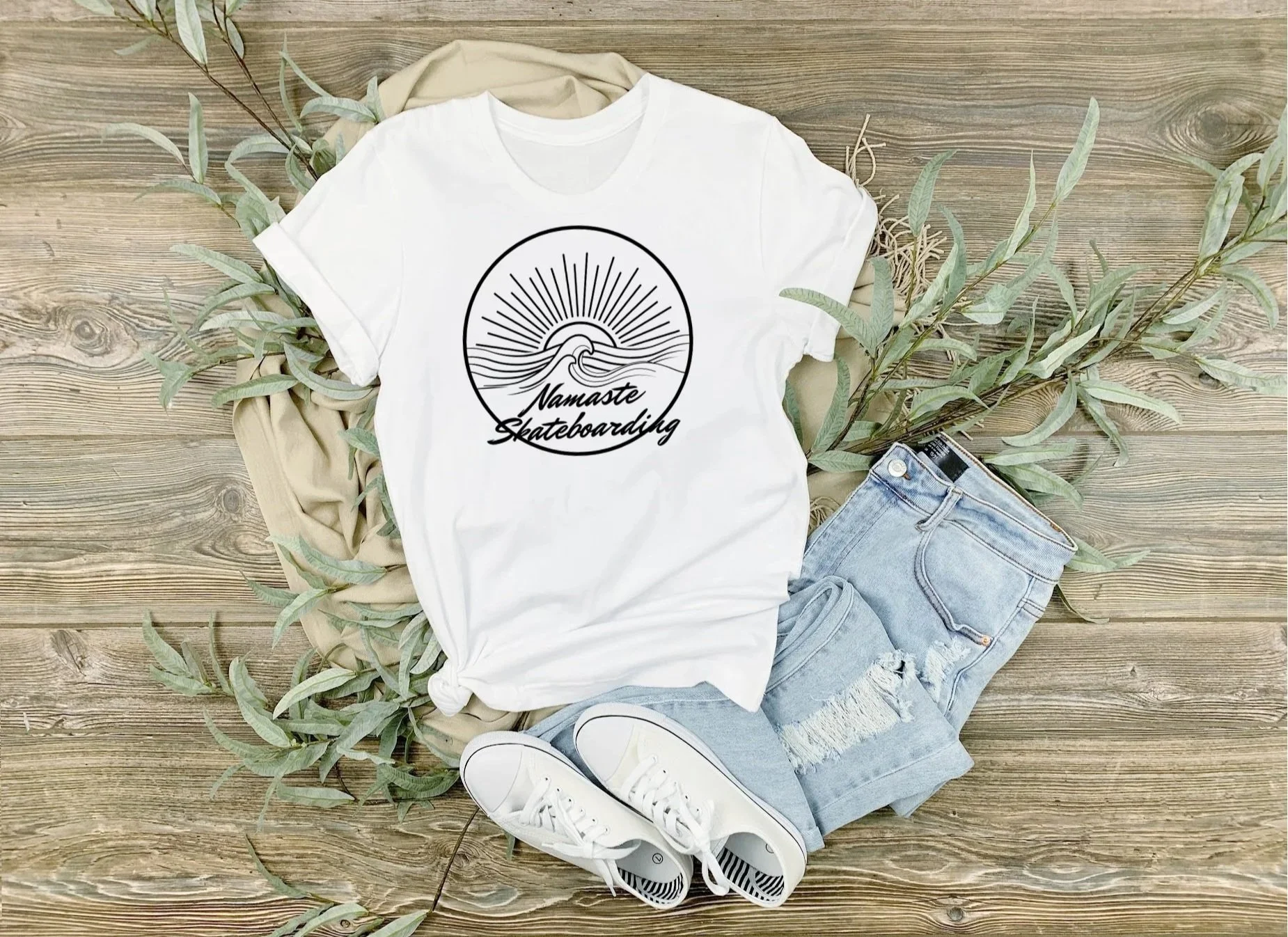 Casual outfit laid out on a wooden floor; white T-shirt with 'Namaste Skateboarding' graphic, light blue ripped denim shorts, white sneakers, beige foliage and a cloth item.