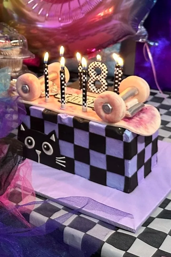 A birthday cake designed to resemble a skateboard, with checkered black and purple sides, a cat face on the front, and five lit candles spelling '18', with a pink 'Happy Birthday' topper and purple balloons in the background.