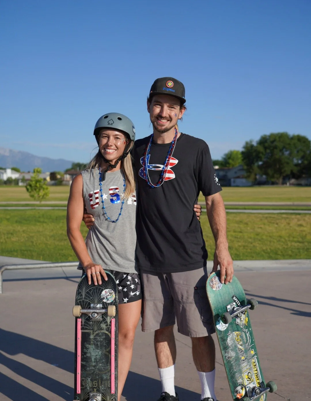 A smiling young woman and man standing together in a park with skateboards, wearing helmets and patriotic-themed accessories, during a sunny day.