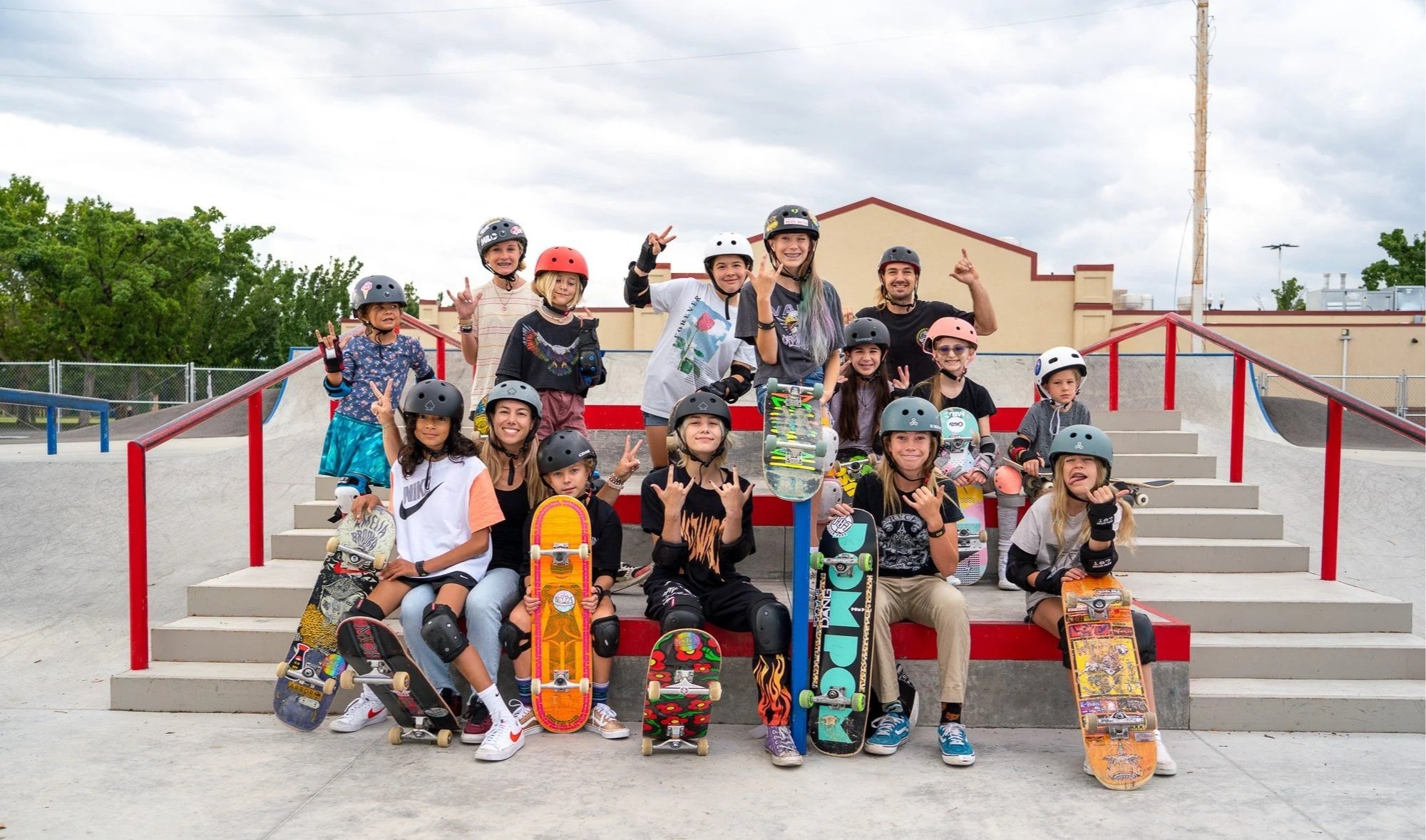 Group of children and teens at a skatepark, wearing helmets, with skateboards and scooters, posing for a photo on the steps under a cloudy sky.