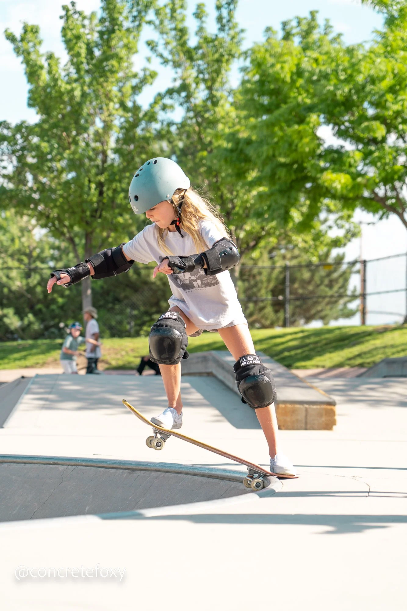 A young girl wearing a helmet, knee pads, and elbow pads skateboarding at a skate park with trees in the background on a sunny day.