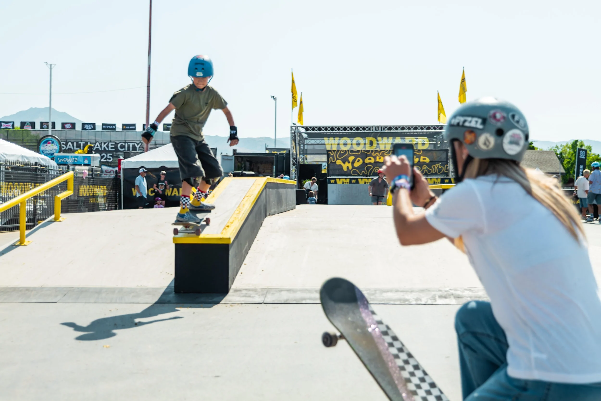 Child in a helmet skateboarding at an outdoor skate park, with a person taking a photo nearby and a banner reading 'Woodward' in the background.