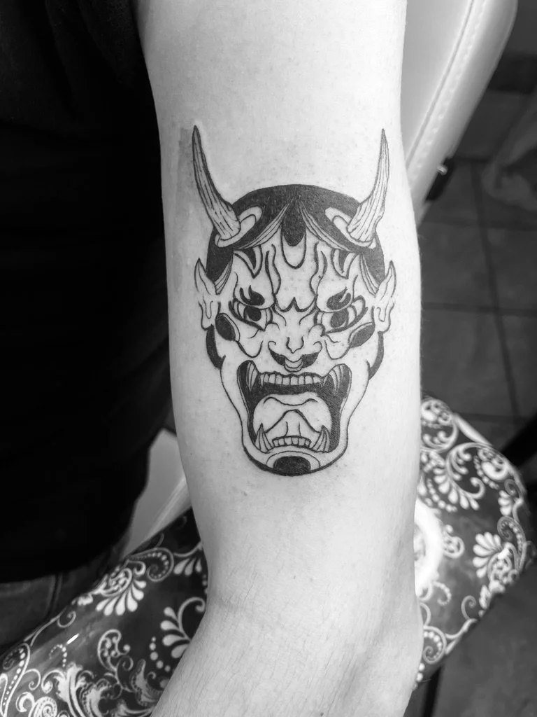 Tattoo of a traditional oni mask with horns, featuring an angry face with sharp teeth and flames around the eyes.