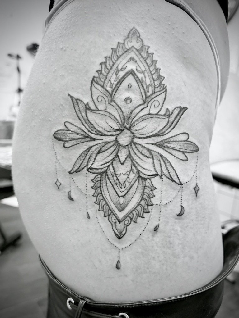 A detailed line art tattoo of a symmetrical pattern with ornamental design elements, floral patterns, and decorative hanging elements on a person's side
