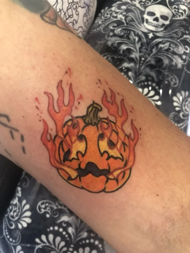 Tattoo of a carved pumpkin with a sinister face, surrounded by flames, on a person's arm.