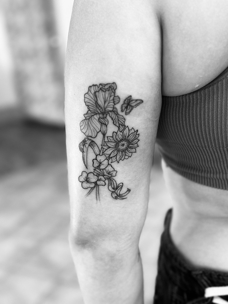 Black and white photo of a person's upper arm showcasing a detailed floral tattoo featuring a sunflower, irises, and smaller flowers with butterflies.
