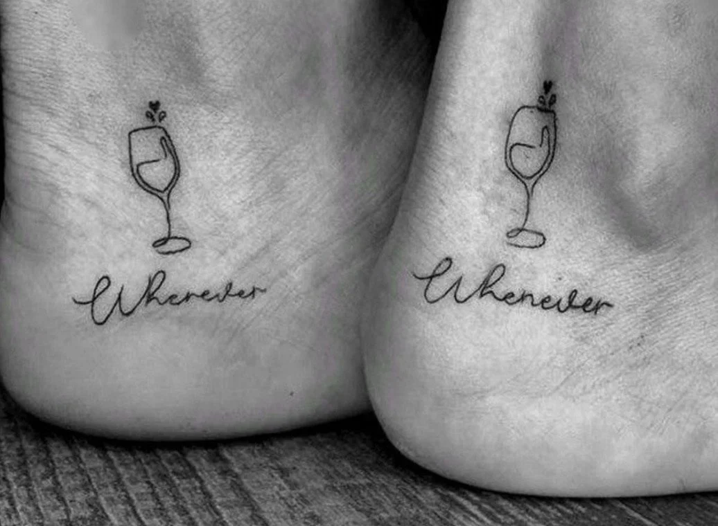 Tattoos of a wine glass with a heart-shaped liquid inside and the word 'Wherever' underneath, on each shoulder blade.