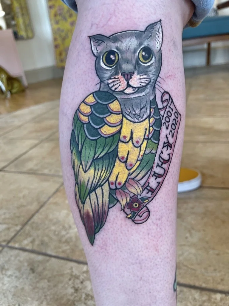 Colorful tattoo of a cat's head combined with a parrot's body on a person's leg, featuring yellow, green, and purple colors, with a banner reading "2007-2022."