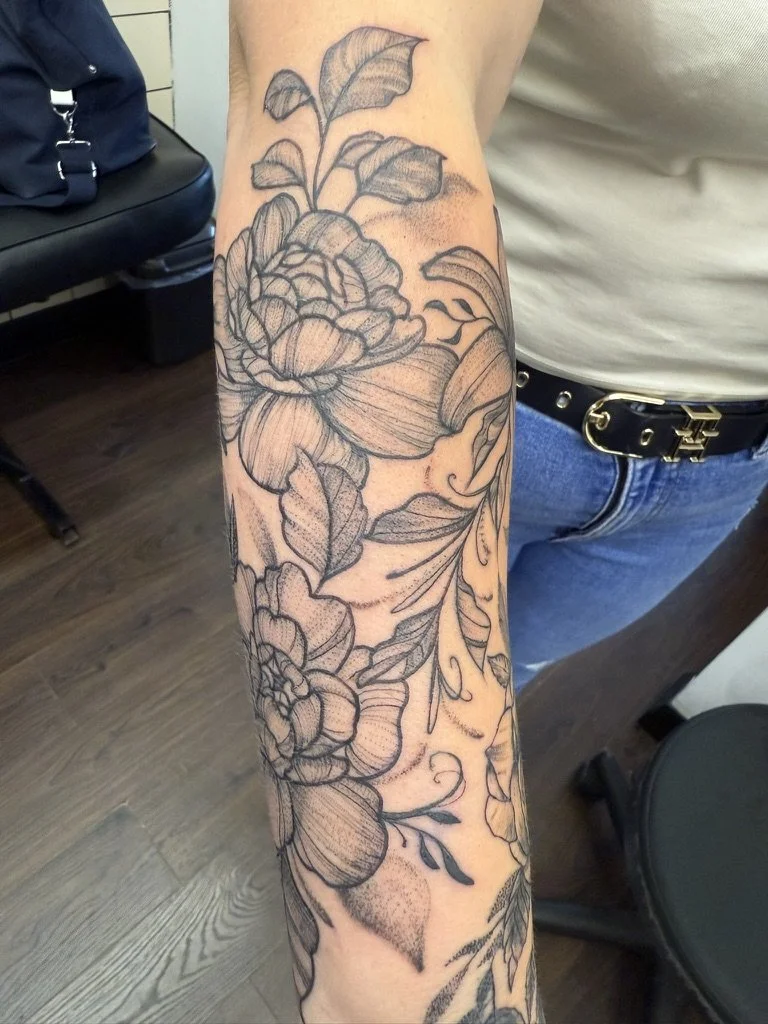 A person's arm with a large black outline fineline floral tattoo in London, featuring detailed flowers and leaves.