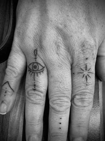 A hand with multiple finger tattoos, including a prominent eye with long eyelashes on the middle finger, decorative lines, and patterns on the other fingers.