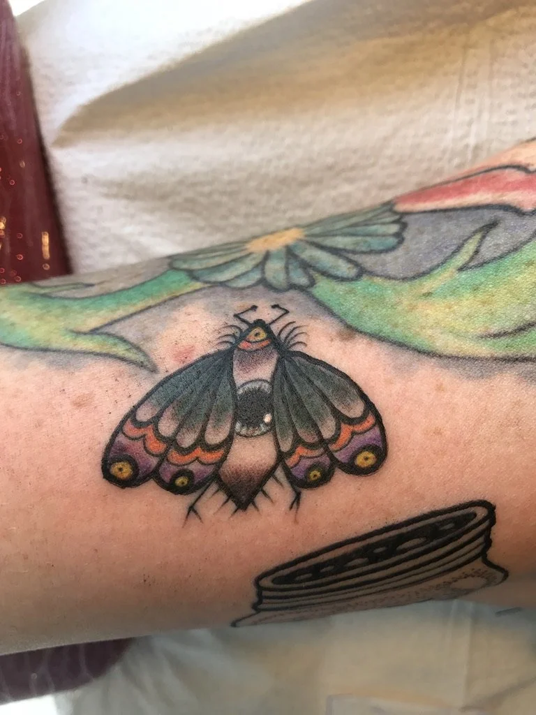 Close-up of a colorful moth tattoo on a person's arm, with other partially visible tattoos around it.