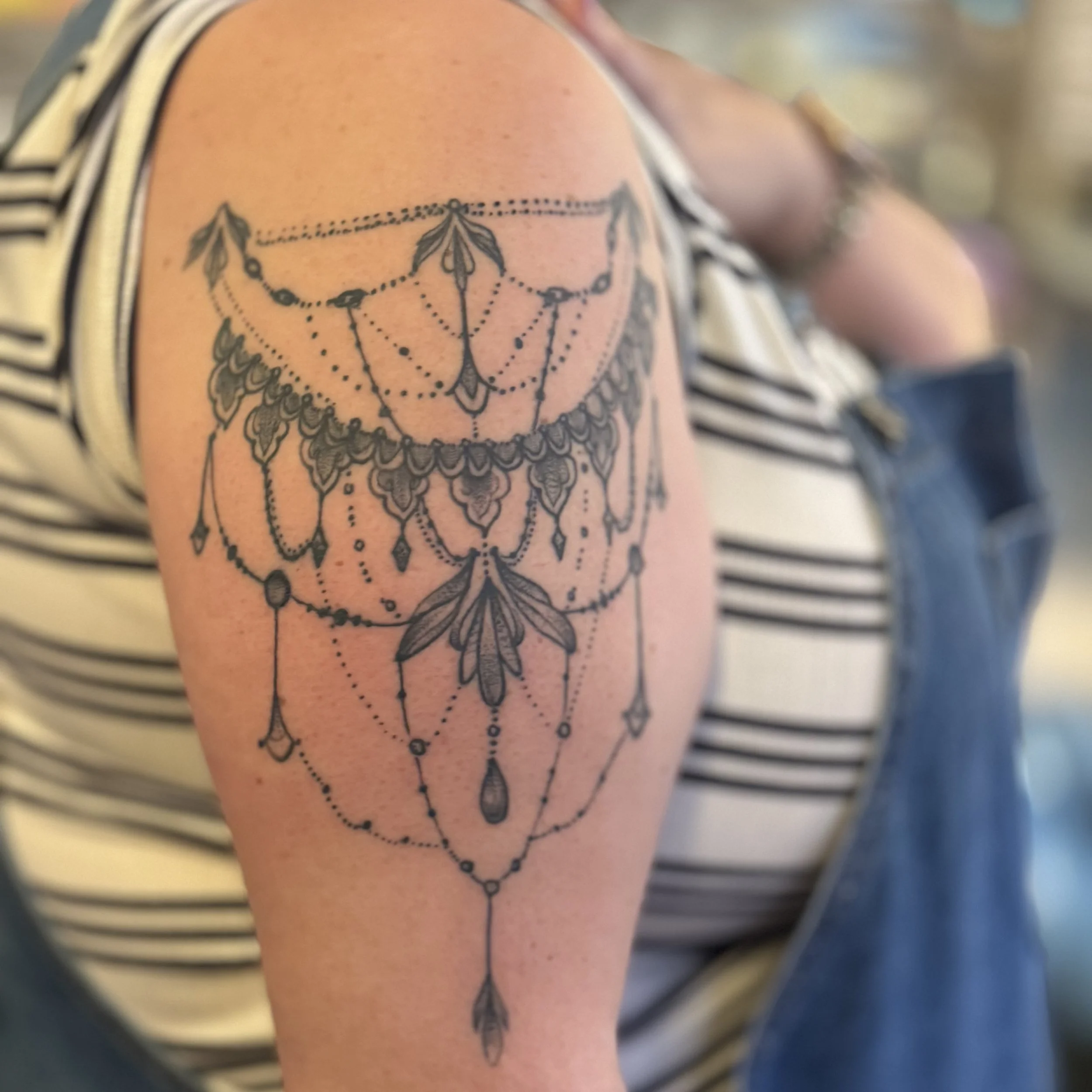 A black line tattoo of chandelier with hanging jewels and beads on a person's upper arm.