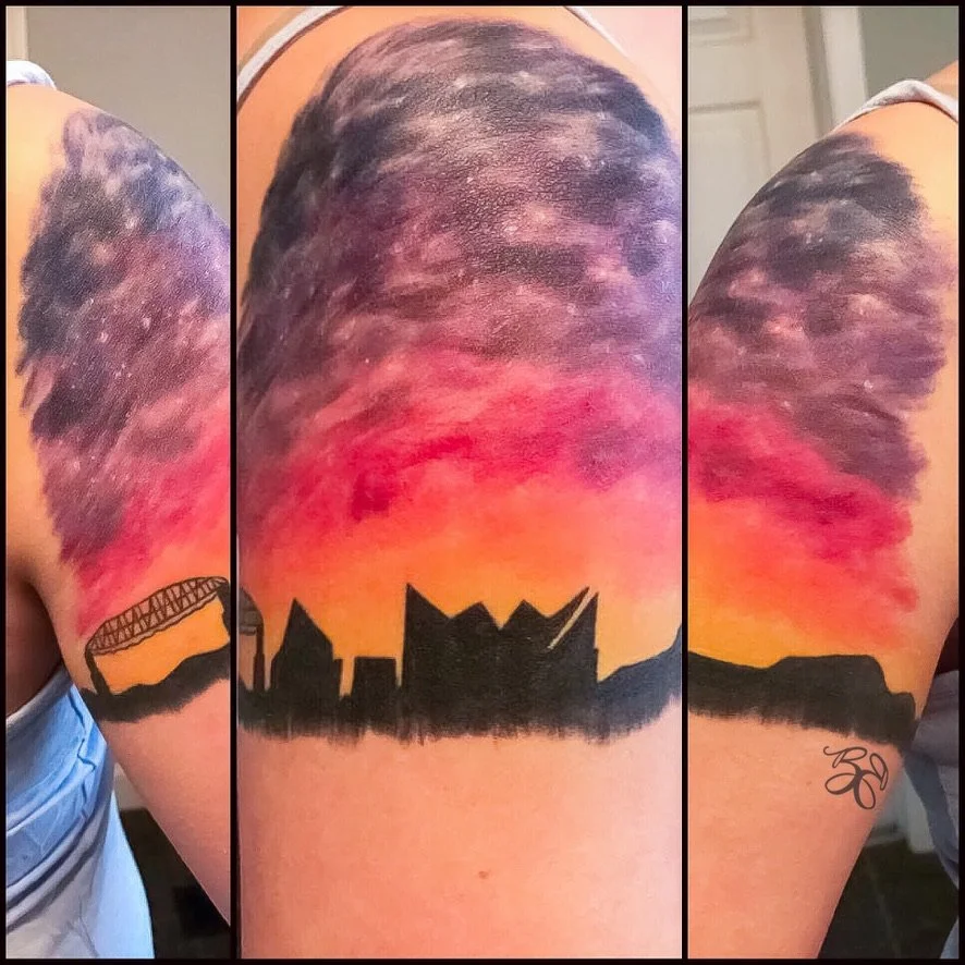 Tattoo of a city skyline with a sunset background, featuring a bridge on the left and a skyline with distinct buildings in the center, on a person's upper arm. The background has vibrant colors of red, orange, pink, and purple, resembling a sunset sk