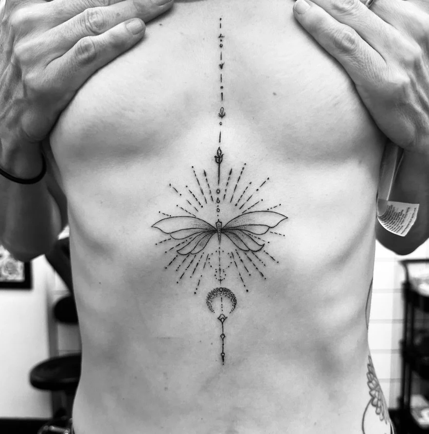 Black and white tattoo of a butterfly with geometric and celestial elements on a person's chest.