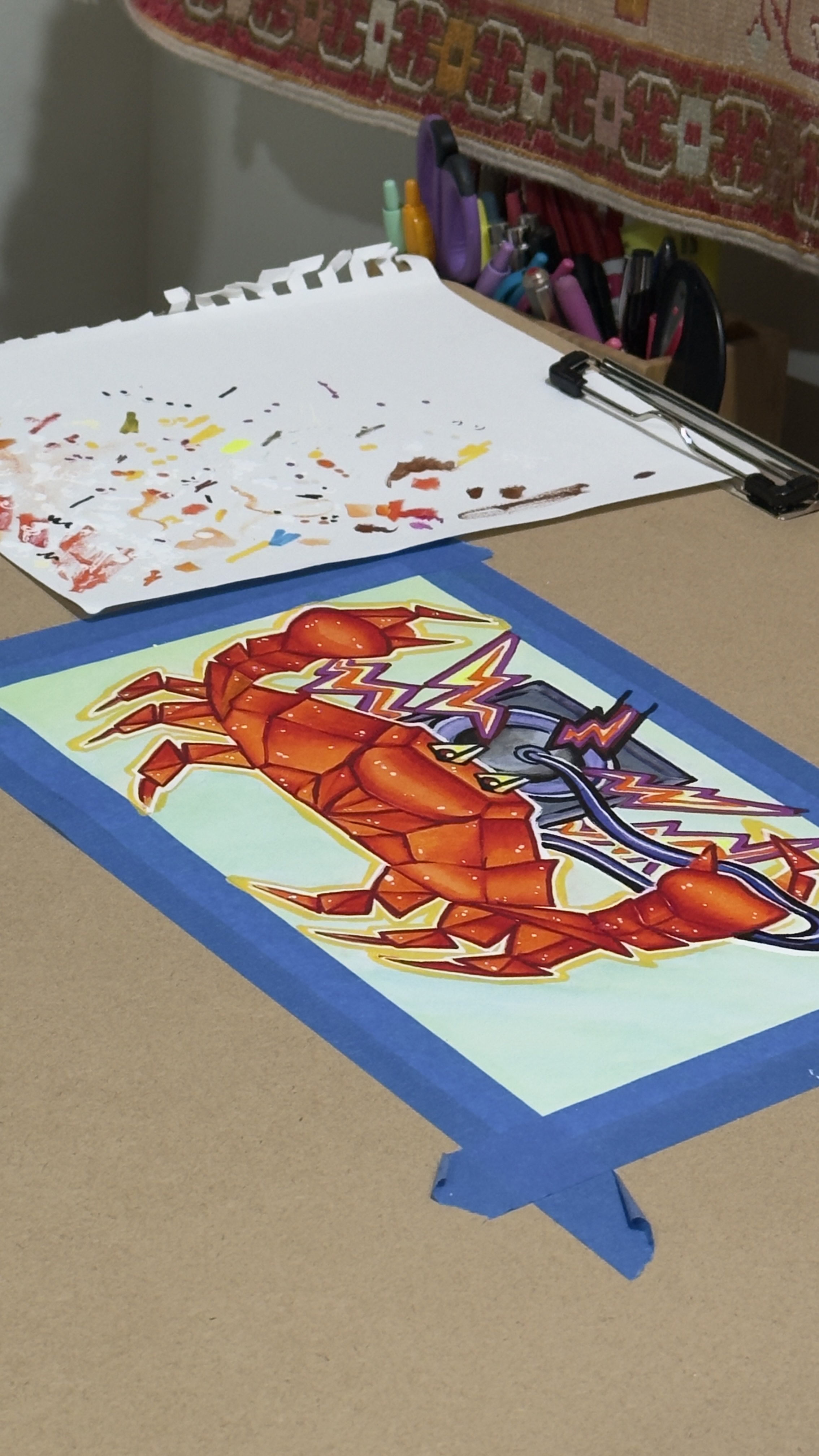 A colorful artwork of a red crab with lightning bolts, taped to a table with blue painter's tape.