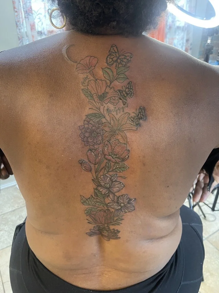 A Black African American person's back with a colorful floral tattoo extending from the upper back to the lower back, featuring flowers, butterflies, and a crescent moon.