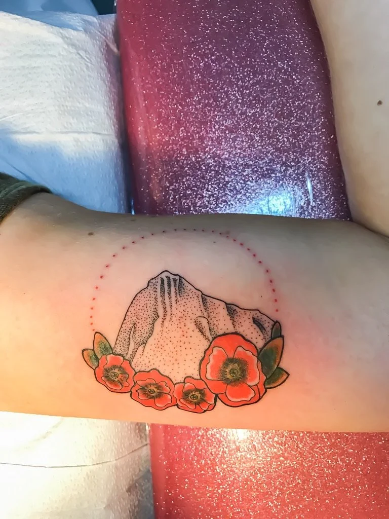 Tattoo of a mountain with five red poppy flowers and green leaves surrounding its base, with dotted lines forming a circle around the mountain and flowers.