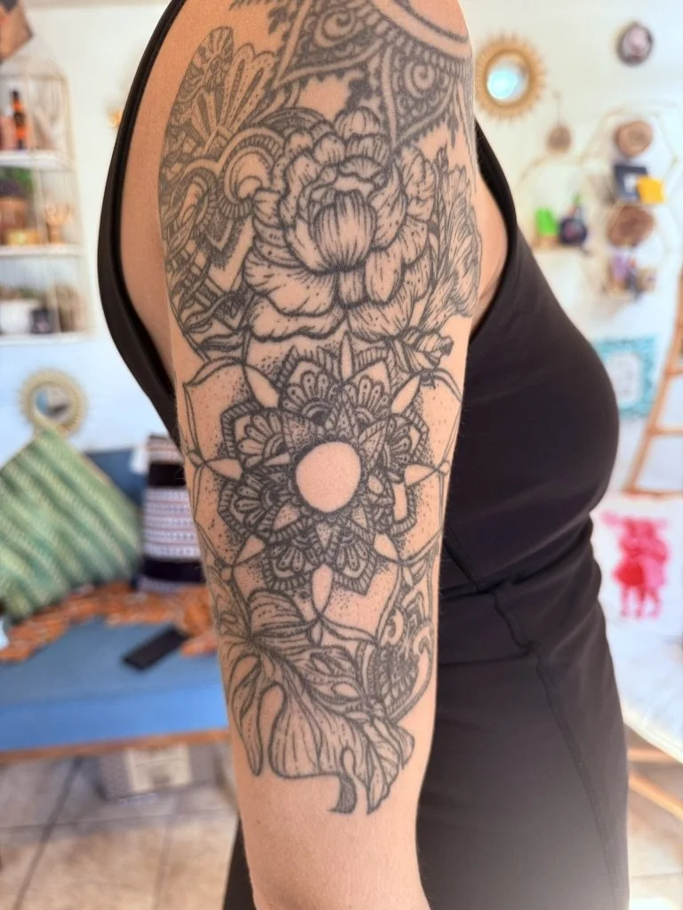 A detailed black and gray floral tattoo on a person's upper arm and shoulder, featuring large roses, mandala patterns, and decorative elements.