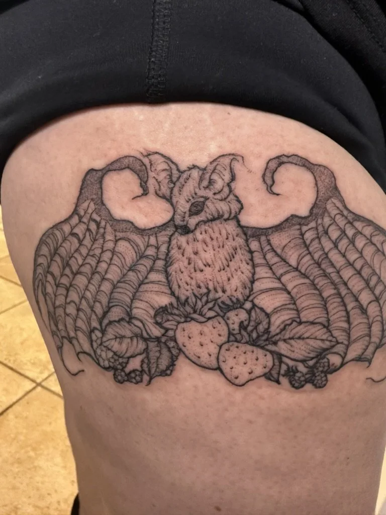 Detailed black and gray tattoo of a bat with wide wings, sitting on strawberries, with intricate line work and shading.